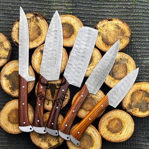 Peut inclure: Ensemble de six couteaux de cuisine avec des manches en bois. Les couteaux ont une finition martel&eacute;e et sont dispos&eacute;s sur une surface en bois. Les couteaux sont &eacute;tiquet&eacute;s "Rick's Metals".
