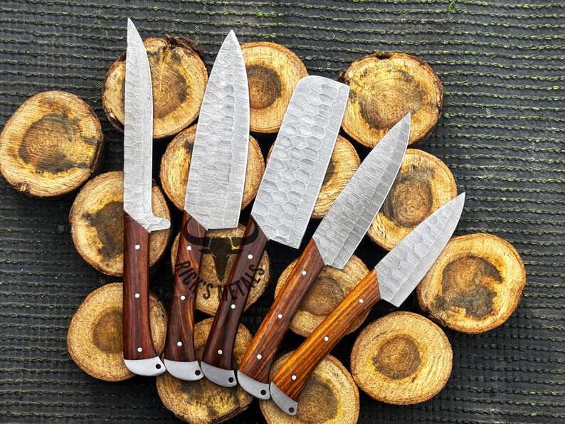 Peut inclure: Ensemble de six couteaux de cuisine avec des manches en bois. Les couteaux ont une finition martel&eacute;e et sont dispos&eacute;s sur une surface en bois. Les couteaux sont &eacute;tiquet&eacute;s "Rick's Metals".