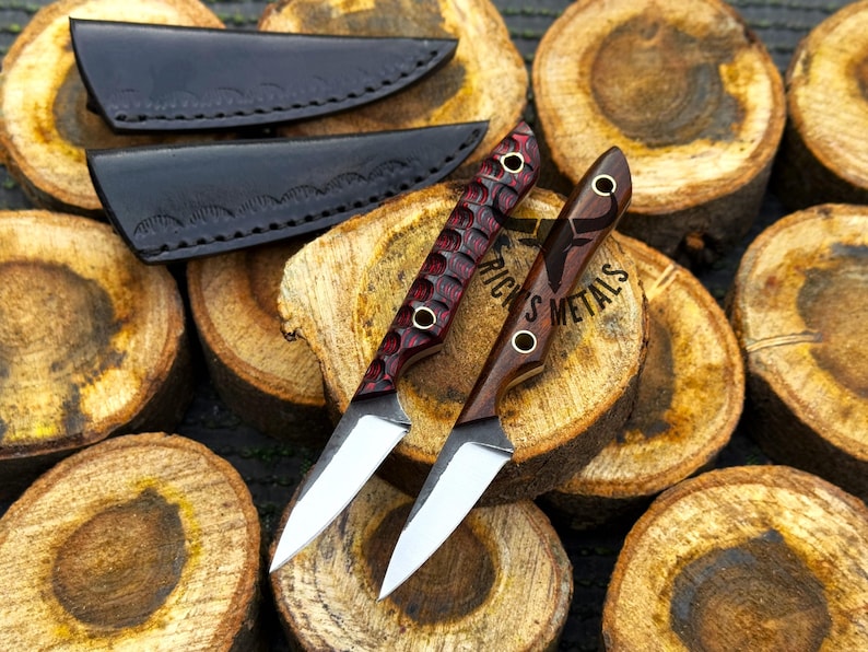 K&ouml;nnte beinhalten: Zwei feststehende Messer mit silbernen Stahlklingen und Holzgriffen. Ein Griff hat eine texturierte, dunkelrote Oberfl&auml;che, w&auml;hrend der andere aus glattem, dunkelbraunem Holz besteht. Beide haben Messingnieten und zwei schwarze Lederscheiden.