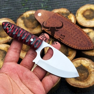 May include: A red and black handled knife with a silver blade and a finger hole in the blade. The knife is held in a hand and is resting on a piece of wood.