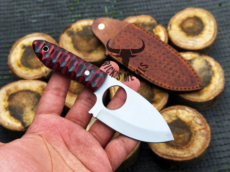 May include: A red and black handled knife with a silver blade and a finger hole in the blade. The knife is held in a hand and is resting on a piece of wood.