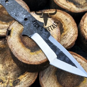 May include: A black and silver knife with a partially polished blade. The knife is resting on a stack of wooden rounds. The text "Rick's Metals" is visible on the knife.