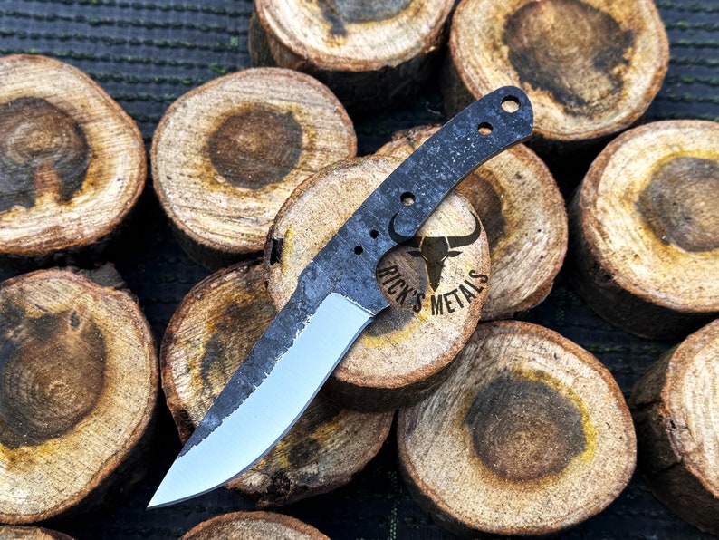 May include: A black and silver knife with a textured blade and a black handle. The knife is lying on a pile of wooden rounds. The knife has the text "Rick's Metals" on the handle.