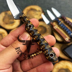 May include: A small knife with a patterned handle in black and gold, featuring a silver blade. The handle has a wavy design with two circular holes. Other knives with different handle colors are in the background. The logo "RICK'S METALS" is also visible.