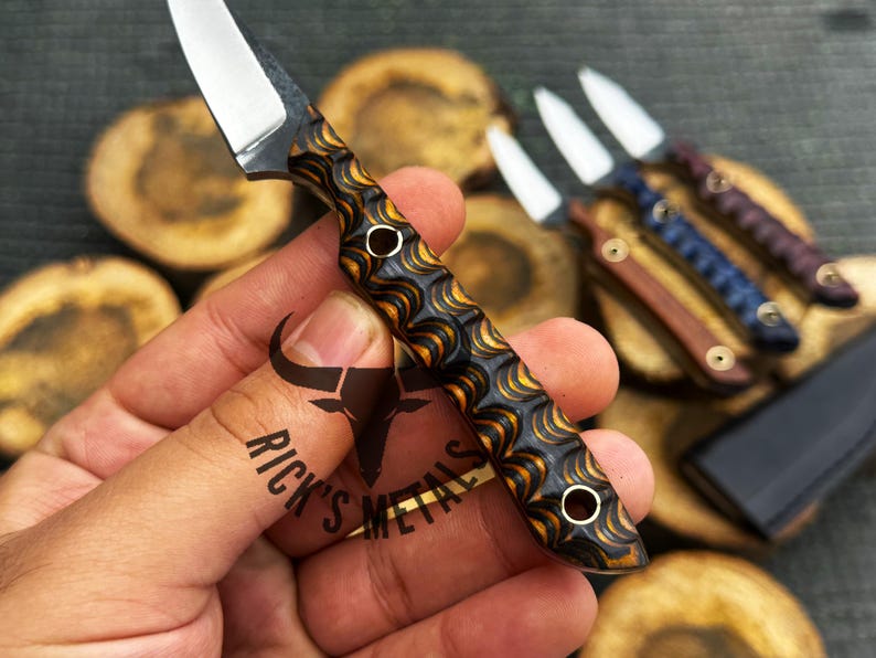 May include: A small knife with a patterned handle in black and gold, featuring a silver blade. The handle has a wavy design with two circular holes. Other knives with different handle colors are in the background. The logo "RICK'S METALS" is also visible.