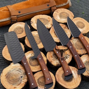 Peut inclure: Cinq couteaux de cuisine noirs avec des manches en bois sont dispos&eacute;s sur une surface en bois. Les couteaux sont dispos&eacute;s en rang&eacute;e, les manches pointant vers le spectateur. Les couteaux sont de tailles et de formes diff&eacute;rentes, et ils pr&eacute;sentent une vari&eacute;t&eacute; de motifs sur leurs lames. Les couteaux sont tous tranchants et ont une finition polie. Les couteaux sont dispos&eacute;s sur une surface en bois, qui est recouverte d'un rouleau en cuir brun. Le rouleau est enroul&eacute; et attach&eacute; avec une sangle en cuir brun. Le rouleau est &eacute;tiquet&eacute; "Ricks Metals".