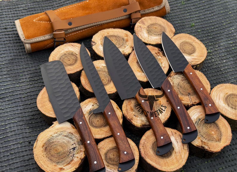 Peut inclure: Cinq couteaux de cuisine noirs avec des manches en bois sont dispos&eacute;s sur une surface en bois. Les couteaux sont dispos&eacute;s en rang&eacute;e, les manches pointant vers le spectateur. Les couteaux sont de tailles et de formes diff&eacute;rentes, et ils pr&eacute;sentent une vari&eacute;t&eacute; de motifs sur leurs lames. Les couteaux sont tous tranchants et ont une finition polie. Les couteaux sont dispos&eacute;s sur une surface en bois, qui est recouverte d'un rouleau en cuir brun. Le rouleau est enroul&eacute; et attach&eacute; avec une sangle en cuir brun. Le rouleau est &eacute;tiquet&eacute; "Ricks Metals".