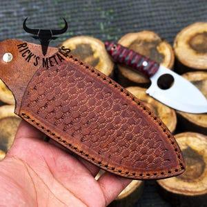 May include: A brown leather sheath with a decorative pattern and stitching. The sheath is designed to hold a small knife with a round hole in the blade. The text "Rick's Metals" is embossed on the sheath.
