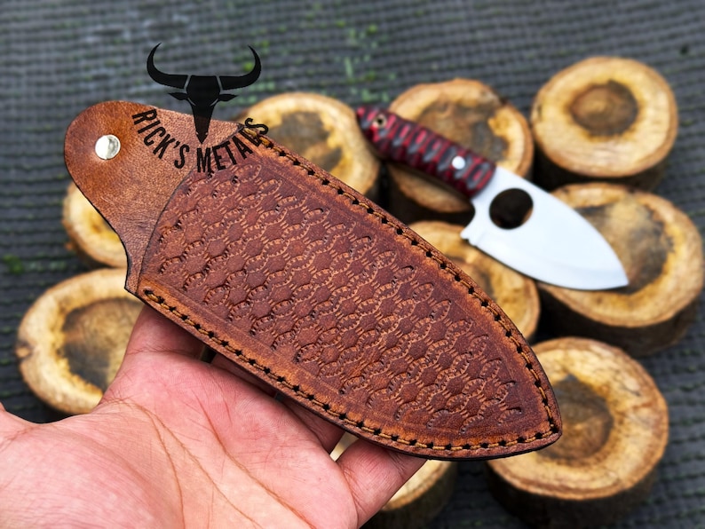 May include: A brown leather sheath with a decorative pattern and stitching. The sheath is designed to hold a small knife with a round hole in the blade. The text "Rick's Metals" is embossed on the sheath.