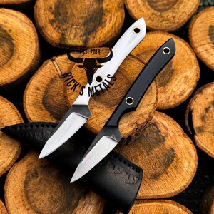 K&ouml;nnte beinhalten: Zwei feststehende Messer mit spitzen Stahlklingen und messingverst&auml;rkten Griff&ouml;ffnungen. Ein Messer hat einen wei&szlig;en Griff, das andere einen schwarzen Griff. Darunter liegt eine schwarze Lederscheide. Der Text RICK'S METALS EST. 2019 ist sichtbar.