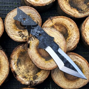 May include: A black and silver knife with a unique shape and a textured finish. The knife is resting on a pile of wooden rounds.