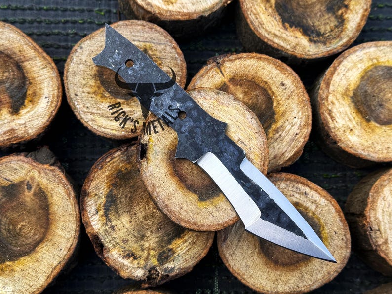 May include: A black and silver knife with a unique shape and a textured finish. The knife is resting on a pile of wooden rounds.