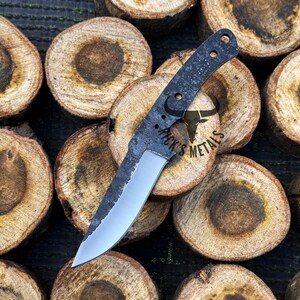 May include: A black and silver knife with a textured handle and a partially sharpened blade. The knife is laying on top of several wooden rounds. The handle has the text "Rick's Metals" on it.