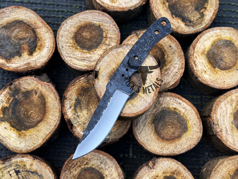 May include: A black and silver knife with a textured handle and a partially sharpened blade. The knife is laying on top of several wooden rounds. The handle has the text "Rick's Metals" on it.