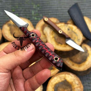 K&ouml;nnte beinhalten: Ein kleines Messer mit feststehender Klinge aus silbernem Stahl und einem strukturierten Griff mit rot-schwarzem Muster sowie zwei Messingnieten. Der Griff ist mit einem schwarzen Stier-Logo und dem Text RICK'S METALS versehen. Ein zweites Messer und eine Scheide sind sichtbar.