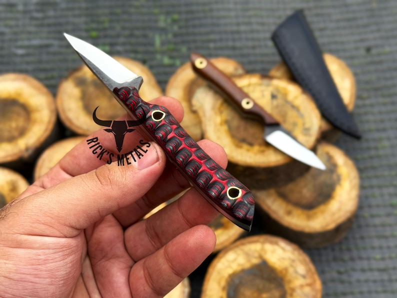 K&ouml;nnte beinhalten: Ein kleines Messer mit feststehender Klinge aus silbernem Stahl und einem strukturierten Griff mit rot-schwarzem Muster sowie zwei Messingnieten. Der Griff ist mit einem schwarzen Stier-Logo und dem Text RICK'S METALS versehen. Ein zweites Messer und eine Scheide sind sichtbar.