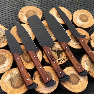 Peut inclure: Cinq couteaux de cuisine noirs avec des manches en bois sont dispos&eacute;s sur une pile de rondins de bois. Les couteaux ont une finition martel&eacute;e et les manches sont d'une couleur brun fonc&eacute;. Les couteaux sont dispos&eacute;s en &eacute;ventail, les lames pointant vers l'ext&eacute;rieur. Le texte "Rick's Metals" est visible sur le manche de l'un des couteaux.