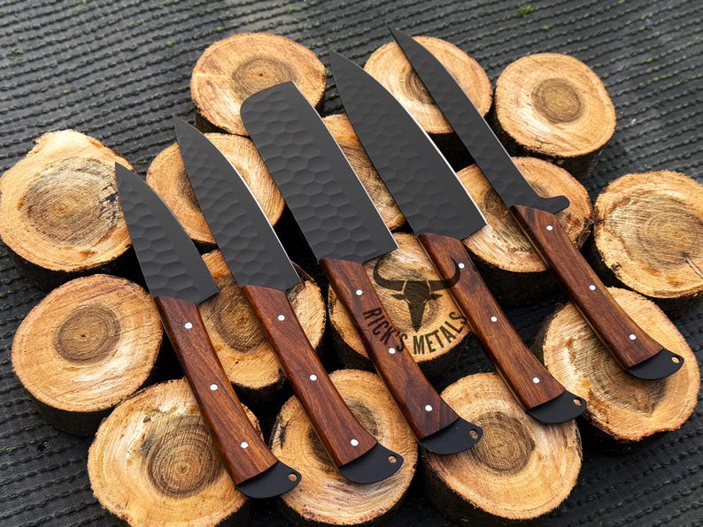 Peut inclure: Cinq couteaux de cuisine noirs avec des manches en bois sont dispos&eacute;s sur une pile de rondins de bois. Les couteaux ont une finition martel&eacute;e et les manches sont d'une couleur brun fonc&eacute;. Les couteaux sont dispos&eacute;s en &eacute;ventail, les lames pointant vers l'ext&eacute;rieur. Le texte "Rick's Metals" est visible sur le manche de l'un des couteaux.