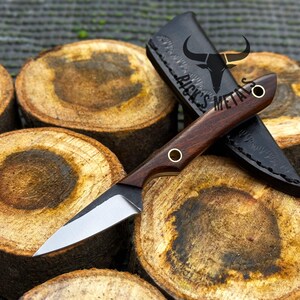 K&ouml;nnte beinhalten: Ein feststehendes Messer mit einer silbernen Stahlklinge und einem dunkelbraunen Holzgriff mit zwei Messingnieten. Das Messer wird von einer schwarzen Lederscheide begleitet, die mit dem Text RICK'S METALS und einem Stierkopf-Logo gepr&auml;gt ist.