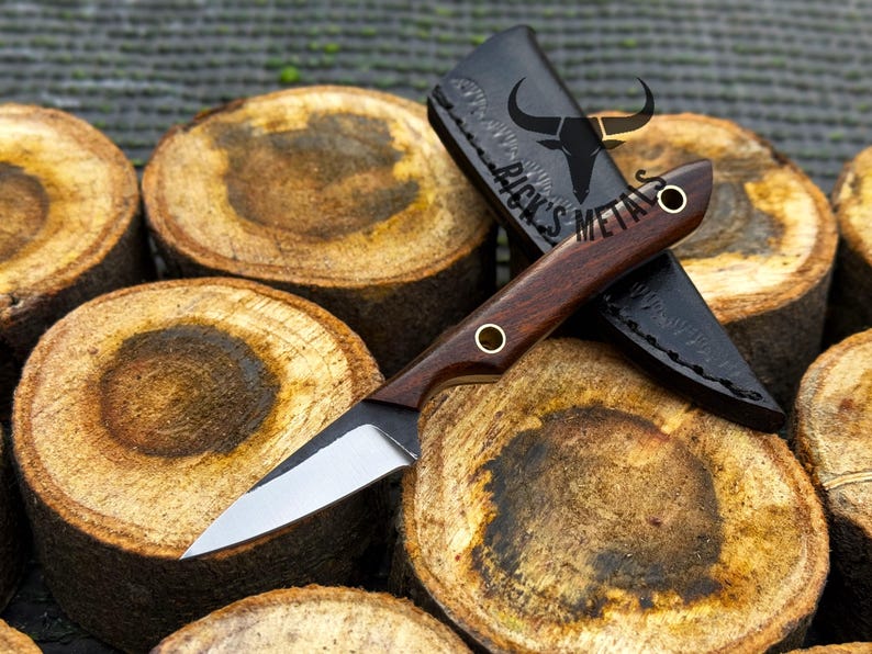 K&ouml;nnte beinhalten: Ein feststehendes Messer mit einer silbernen Stahlklinge und einem dunkelbraunen Holzgriff mit zwei Messingnieten. Das Messer wird von einer schwarzen Lederscheide begleitet, die mit dem Text RICK'S METALS und einem Stierkopf-Logo gepr&auml;gt ist.