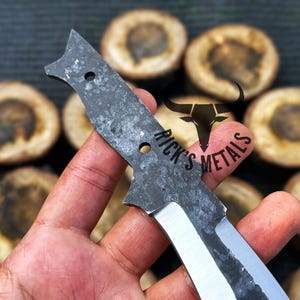 May include: A hand holds a black and silver knife blade with a rough, textured surface. The blade has two holes and the text "Rick's Metals" is visible on the blade.