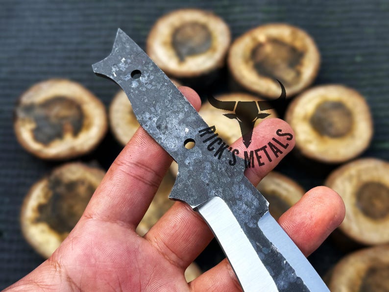 May include: A hand holds a black and silver knife blade with a rough, textured surface. The blade has two holes and the text "Rick's Metals" is visible on the blade.