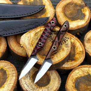 K&ouml;nnte beinhalten: Zwei kleine Messer mit feststehender Klinge, silbernen Stahlklingen und Holzgriffen. Ein Griff hat ein strukturiertes rot-schwarzes Muster, der andere besteht aus glattem, dunkelbraunem Holz mit dem Schriftzug RICK'S METALS. Zwei schwarze Lederscheiden liegen bei.