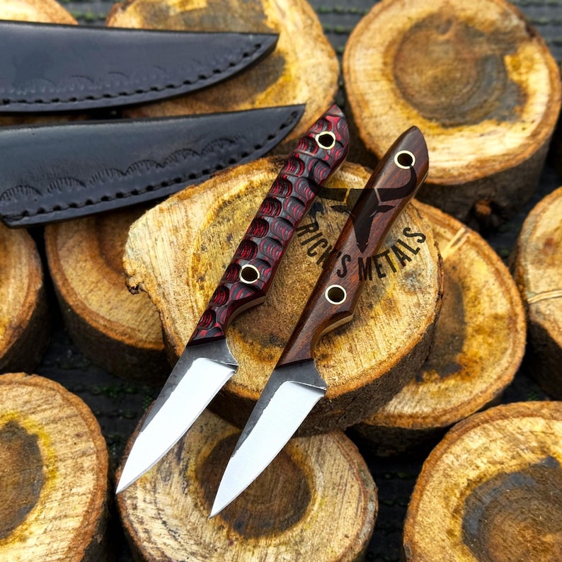 K&ouml;nnte beinhalten: Zwei kleine Messer mit feststehender Klinge, silbernen Stahlklingen und Holzgriffen. Ein Griff hat ein strukturiertes rot-schwarzes Muster, der andere besteht aus glattem, dunkelbraunem Holz mit dem Schriftzug RICK'S METALS. Zwei schwarze Lederscheiden liegen bei.