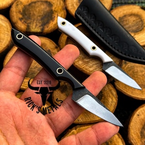 K&ouml;nnte beinhalten: Ein kleines Messer mit feststehender Klinge, schwarzem Griff und zwei Messingnieten sowie einer silbernen Stahlklinge. Ein zweites Messer mit wei&szlig;em Griff und eine schwarze Lederscheide sind zu sehen. Der Text RICK'S METALS EST. 2019 steht auf dem Griff.