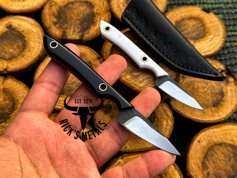 K&ouml;nnte beinhalten: Ein kleines Messer mit feststehender Klinge, schwarzem Griff und zwei Messingnieten sowie einer silbernen Stahlklinge. Ein zweites Messer mit wei&szlig;em Griff und eine schwarze Lederscheide sind zu sehen. Der Text RICK'S METALS EST. 2019 steht auf dem Griff.