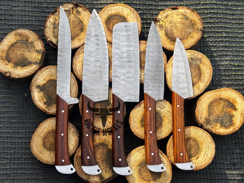 Peut inclure: Six couteaux de cuisine avec des manches en bois et une finition martel&eacute;e. Les couteaux sont dispos&eacute;s sur un fond de rondins de bois. Le texte "Rick's Metals" est visible sur les lames.