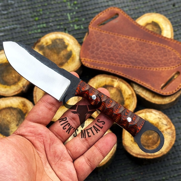 Puede incluir: Un pequeño cuchillo hecho a mano con mango negro y hoja plateada. El mango tiene un patrón rojo y negro y un pequeño agujero en el extremo. El cuchillo está siendo sostenido en una mano.