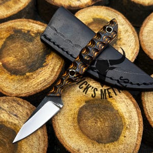 May include: A small, handcrafted knife with a patterned handle and a leather sheath. The handle features a brown and orange wave design. The sheath is black with a decorative edge and a bull graphic. The knife and sheath rest on a wooden surface with the text "RICK'S MEAT".