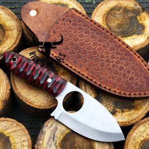 May include: A small, silver knife with a red and black handle. The knife has a finger hole in the blade and is sitting on a pile of wooden rounds. The knife comes with a brown leather sheath with a decorative pattern.