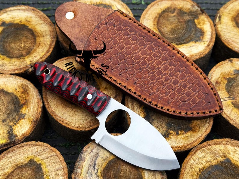 May include: A small, silver knife with a red and black handle. The knife has a finger hole in the blade and is sitting on a pile of wooden rounds. The knife comes with a brown leather sheath with a decorative pattern.