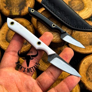 K&ouml;nnte beinhalten: Ein kleines Messer mit feststehender Klinge, wei&szlig;em Griff und zwei Messingnieten sowie einer scharfen Stahlklinge. Ein zweites identisches Messer mit schwarzem Griff und schwarzer Lederscheide ist im Hintergrund zu sehen. Der Text Rick's Metals Est 2019 ist vorhanden.