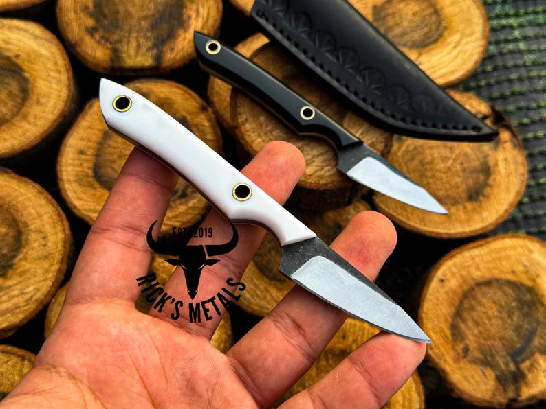 K&ouml;nnte beinhalten: Ein kleines Messer mit feststehender Klinge, wei&szlig;em Griff und zwei Messingnieten sowie einer scharfen Stahlklinge. Ein zweites identisches Messer mit schwarzem Griff und schwarzer Lederscheide ist im Hintergrund zu sehen. Der Text Rick's Metals Est 2019 ist vorhanden.