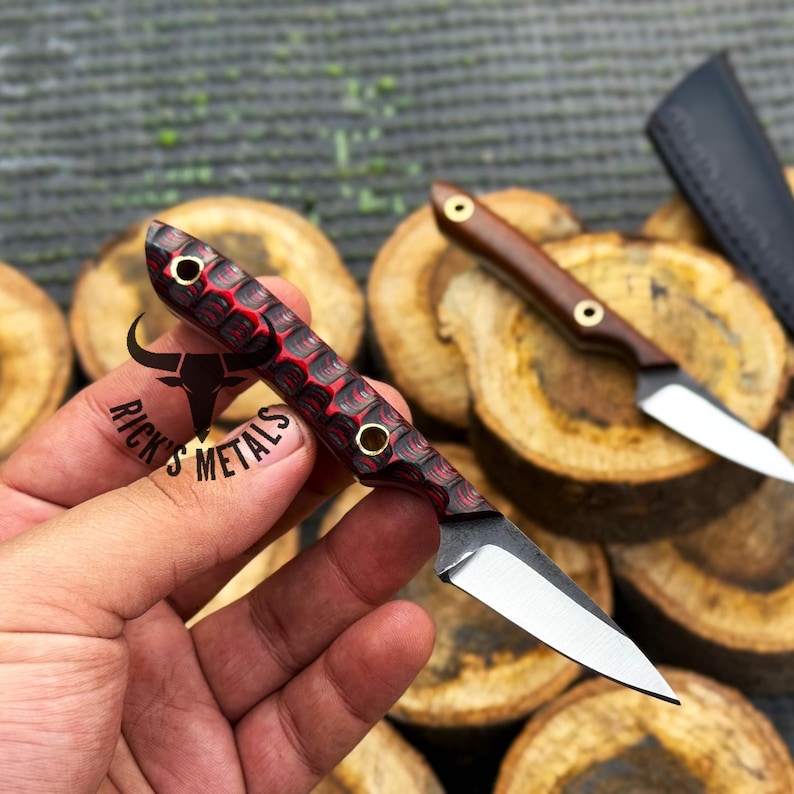 K&ouml;nnte beinhalten: Ein kleines Messer mit feststehender Klinge und einem strukturierten rot-schwarzen Griff, der zwei Messingnieten und eine Fangriemen&ouml;se aufweist. Die Klinge ist aus silberfarbenem Stahl. Der Griff tr&auml;gt ein Stier-Logo und den Text RICK'S METALS.