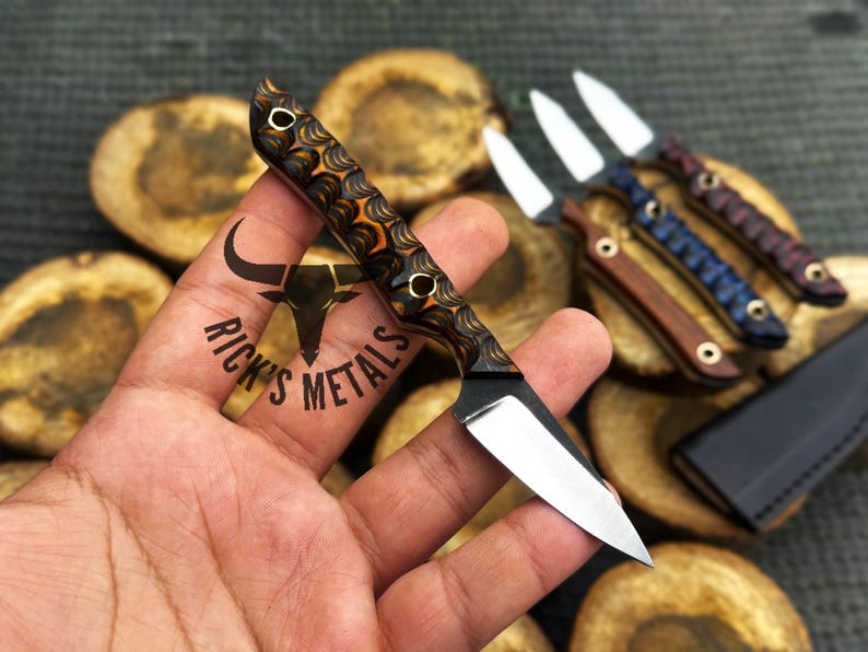 K&ouml;nnte beinhalten: Ein kleines Messer mit feststehender Klinge aus poliertem silberfarbenem Stahl und einem konturierten Griff mit einem geschichteten orangefarbenen, schwarzen und braunen Muster. Der Griff hat zwei messingfarbene Nieten. Der Text Rick's Metals ist sichtbar.