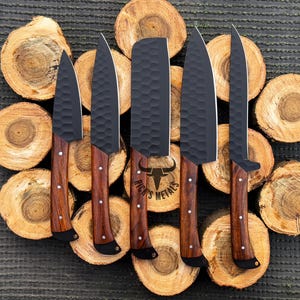 Peut inclure: Ensemble de cinq couteaux noirs avec des manches en bois. Les couteaux ont une finition textur&eacute;e et sont dispos&eacute;s sur une surface en bois. Le plus grand couteau porte l'inscription "Rick's Metals" sur la lame.