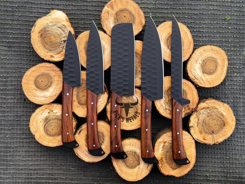 Peut inclure: Ensemble de cinq couteaux noirs avec des manches en bois. Les couteaux ont une finition textur&eacute;e et sont dispos&eacute;s sur une surface en bois. Le plus grand couteau porte l'inscription "Rick's Metals" sur la lame.
