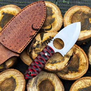 May include: A red and black patterned knife with a finger hole in the blade and a leather sheath. The knife is resting on a pile of wooden rounds.