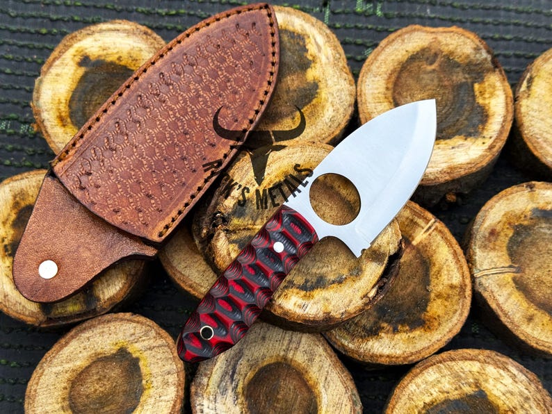 May include: A red and black patterned knife with a finger hole in the blade and a leather sheath. The knife is resting on a pile of wooden rounds.