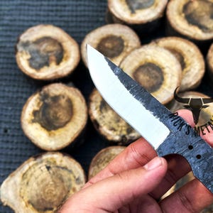 May include: A hand holds a black and white knife with a partially polished blade. The knife has a black handle with three holes and the text "Rick's Metals" is visible on the handle.