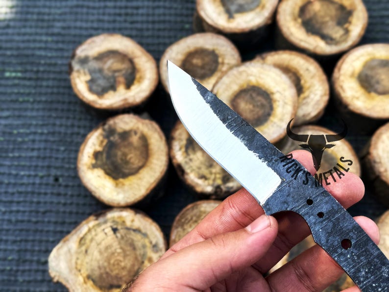 May include: A hand holds a black and white knife with a partially polished blade. The knife has a black handle with three holes and the text "Rick's Metals" is visible on the handle.
