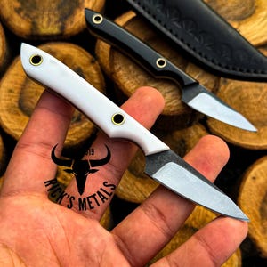 May include: A small fixed-blade knife with a white handle, two brass rivets, and a silver-colored steel blade. A second black-handled knife and a black leather sheath are visible in the background. The text Rick's Metals is printed on the blade.