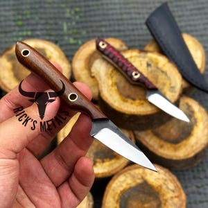 May include: Handmade small knives with wooden handles and metal blades. One knife is held in a hand, while another rests on a wooden surface. The handles are brown and red, with brass accents. The text "Rick's Metals" is visible.