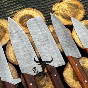 Peut inclure: Cinq couteaux de cuisine avec des manches en bois et une finition en acier martel&eacute;. Les couteaux sont dispos&eacute;s sur une surface en bois. Les couteaux sont de tailles diff&eacute;rentes et pr&eacute;sentent un motif unique sur la lame. Le texte "Rick's Metals" est visible sur la lame de l'un des couteaux.