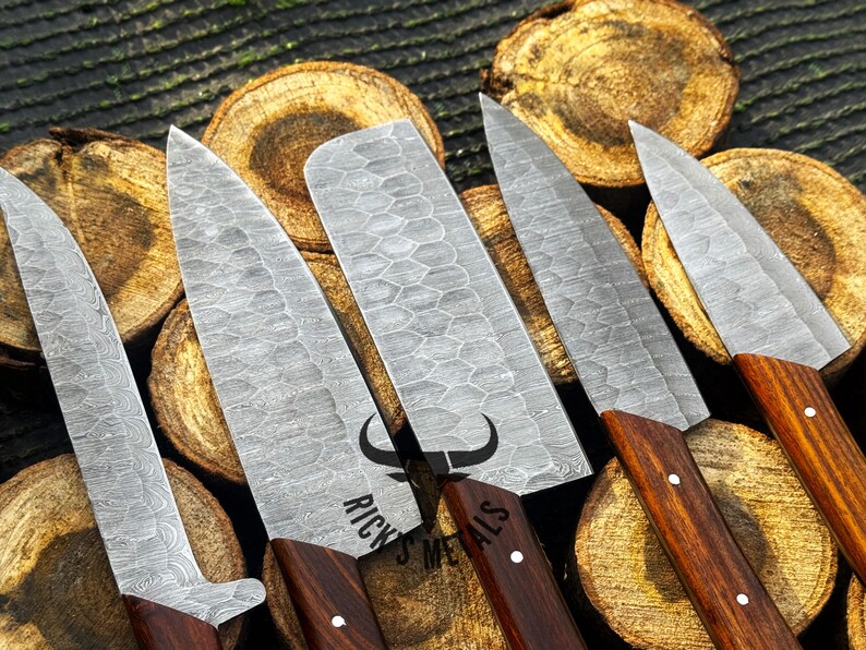 Peut inclure: Cinq couteaux de cuisine avec des manches en bois et une finition en acier martel&eacute;. Les couteaux sont dispos&eacute;s sur une surface en bois. Les couteaux sont de tailles diff&eacute;rentes et pr&eacute;sentent un motif unique sur la lame. Le texte "Rick's Metals" est visible sur la lame de l'un des couteaux.