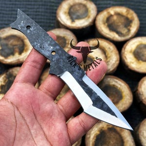 May include: A hand holds a small, black and silver knife with a sharp point. The knife has a textured handle and a partially polished blade. The background is blurred and shows a pile of wooden rounds.
